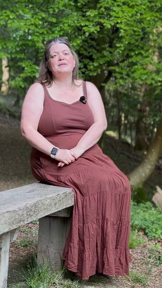 Carly sitting on a wooden bench in Plymbridge Woods. She is wearing a long brown summer dress.